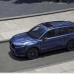 A high-angle view of a dark blue 2026 Honda CR-V Hybrid driving along a paved city street next to a curved sidewalk.