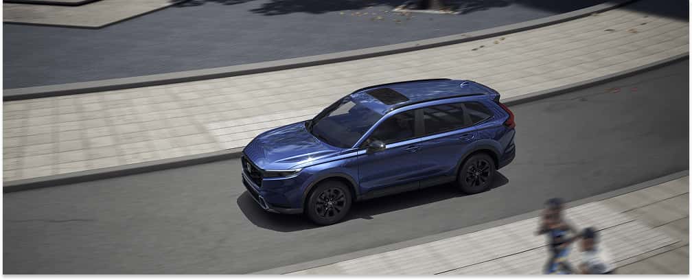 A high-angle view of a dark blue 2026 Honda CR-V Hybrid driving along a paved city street next to a curved sidewalk.