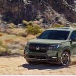 Front three-quarter view of a green 2026 Honda Ridgeline pickup truck driving on a dusty off-road trail.