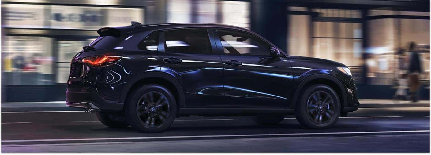 Rear three-quarter view of a dark blue 2027 Honda HR-V driving down an urban city street at night.
