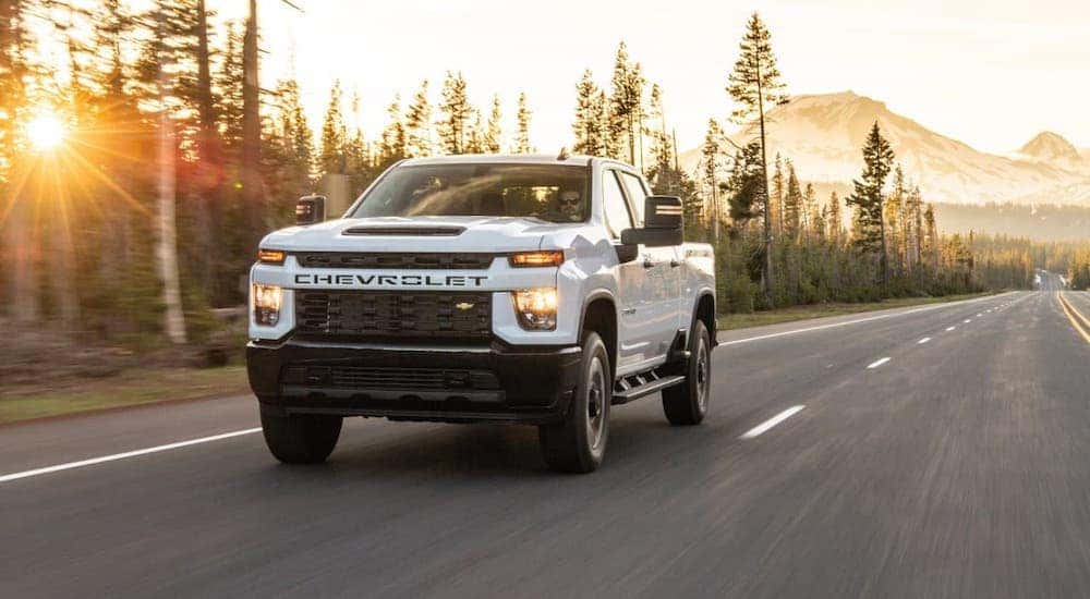 A white 2020 Chevy Silverado 2500HD Custom from a used truck dealer is driving on a highway past evergreen trees.