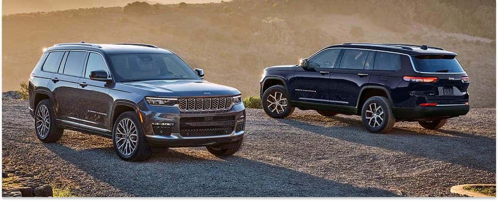 Front three-quarter view of a gray 2026 Jeep Grand Cherokee parked beside a dark blue model on a gravel road.