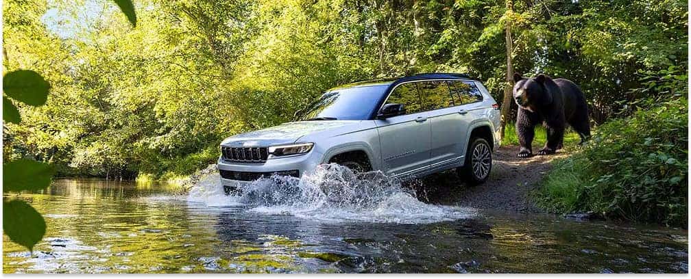 Silver 2026 Jeep Grand Cherokee SUV driving through a shallow river in a forested area.