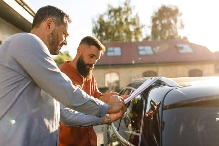 man at used car dealership reviewing papers with employee near car that is for sale