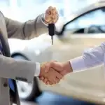 Close-up of two men in business suits shaking hands. One man holds car keys during the handshake. A blurred white car is visible in the background inside a dealership.