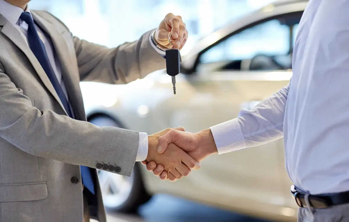 Close-up of two men in business suits shaking hands. One man holds car keys during the handshake. A blurred white car is visible in the background inside a dealership.