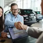 A happy couple sitting at a dealership desk. The smiling man shakes hands with a salesperson across the desk, which has an open laptop on it. Display cars are visible in the background.