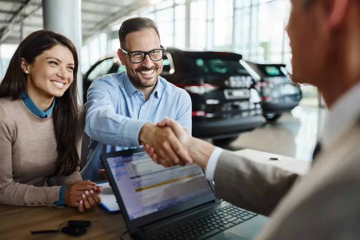 A happy couple sitting at a dealership desk. The smiling man shakes hands with a salesperson across the desk, which has an open laptop on it. Display cars are visible in the background.