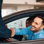 A smiling man sitting in the driver's seat of a car shaking hands with a salesperson outside the vehicle. A smiling woman looks on from the passenger seat.