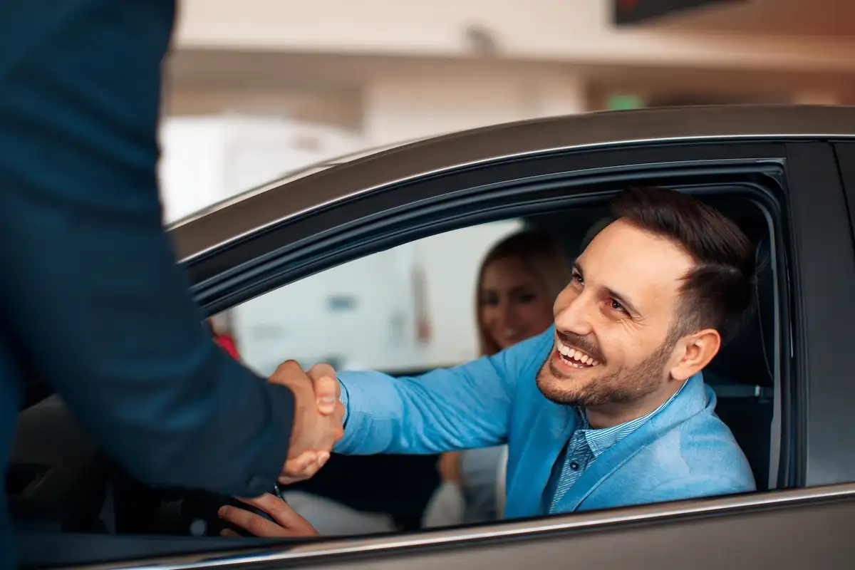 A smiling man sitting in the driver's seat of a car shaking hands with a salesperson outside the vehicle. A smiling woman looks on from the passenger seat.