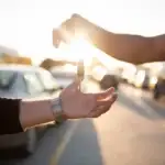 Close-up of a hand dropping car keys into another person's open palm. The sun shines brightly in the background creating a lens flare, with blurred parked cars on the street.