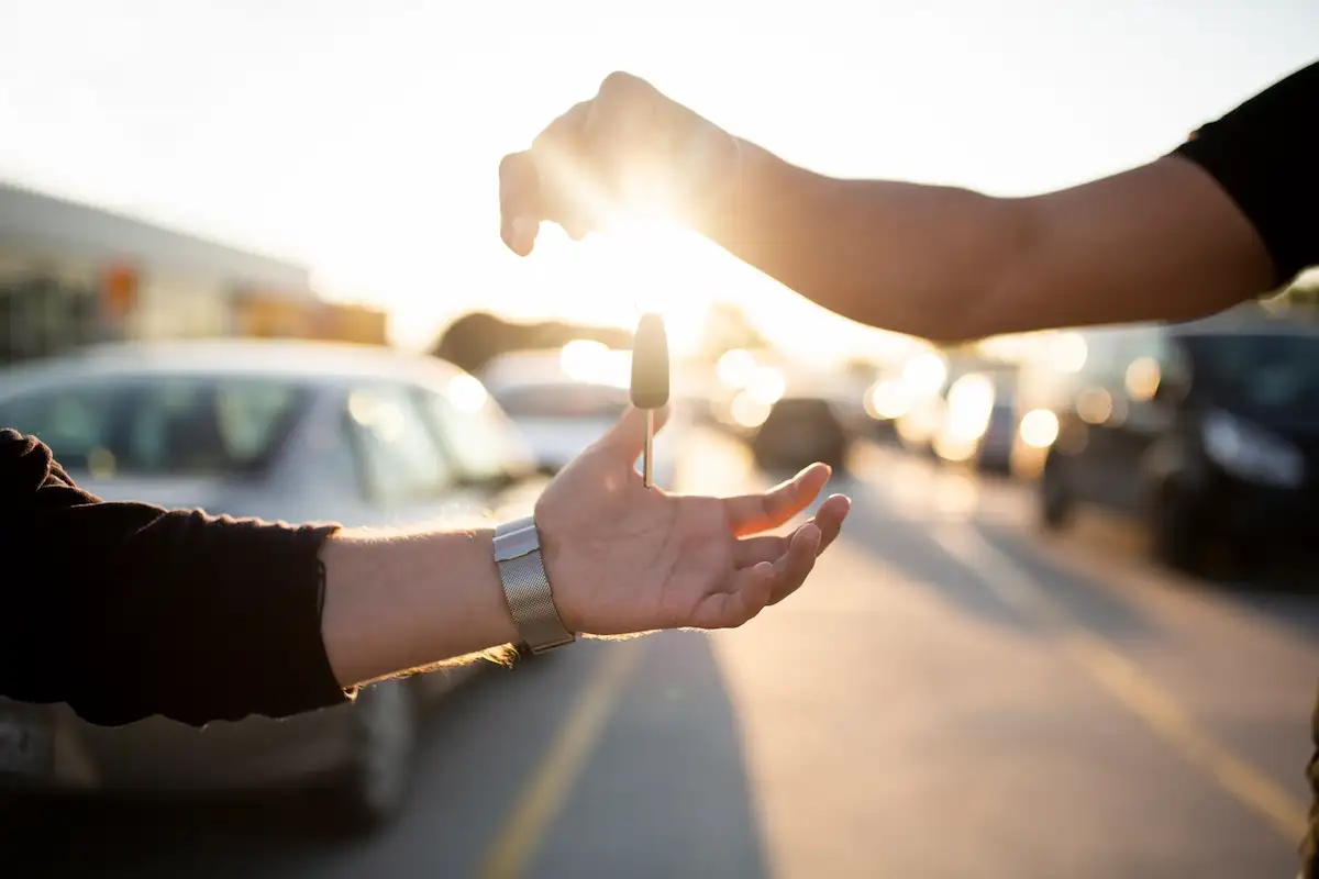 Close-up of a hand dropping car keys into another person's open palm. The sun shines brightly in the background creating a lens flare, with blurred parked cars on the street.