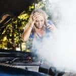 This distraught young woman, stranded after a car breakdown, looks down at her smoking car engine, hand to her head, grimacing in horrified confusion and frustration.
