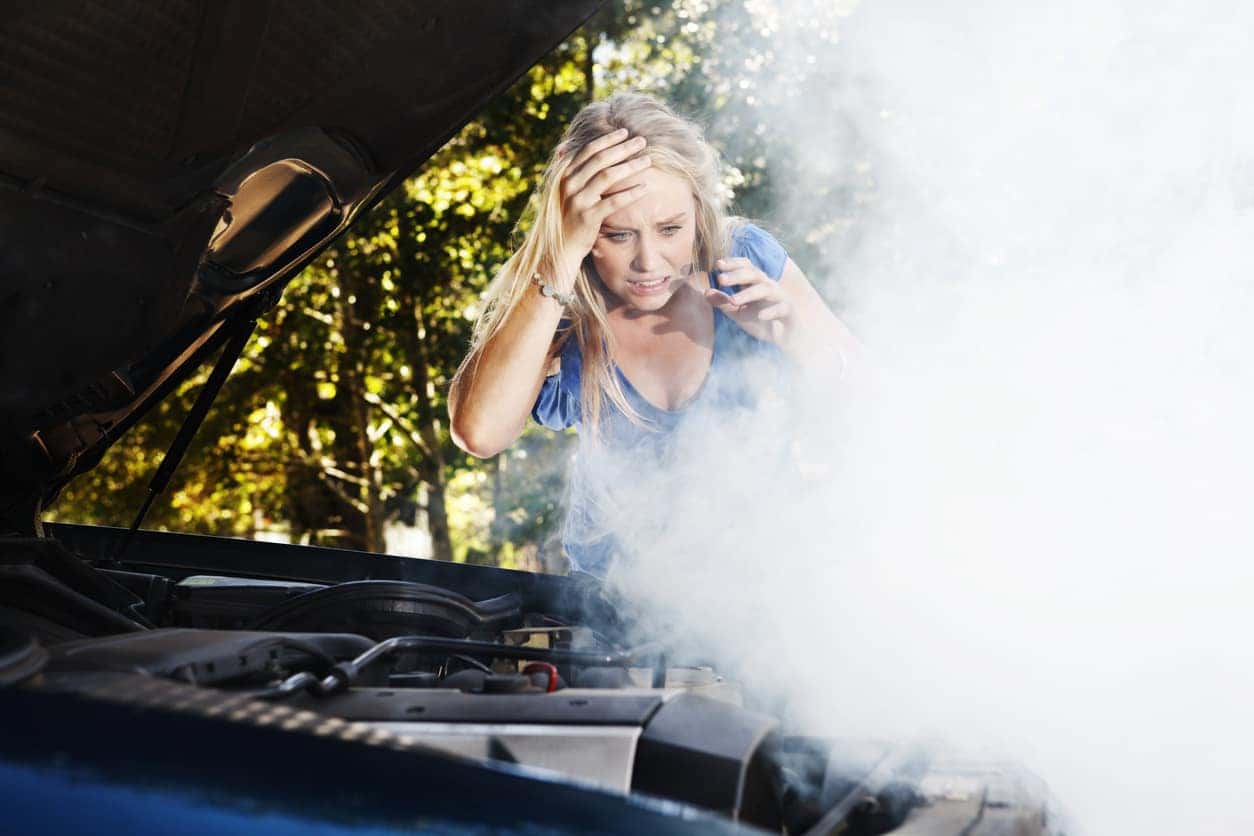 This distraught young woman, stranded after a car breakdown, looks down at her smoking car engine, hand to her head, grimacing in horrified confusion and frustration.