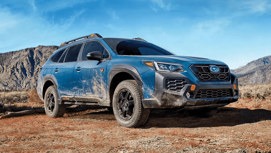 A muddy blue Subaru with rugged black trim and off-road tires is parked on a dirt trail, with dry mountains and a blue sky in the background.
