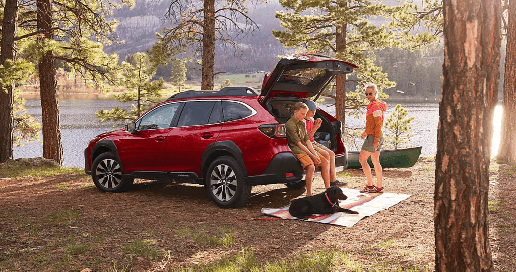 A red Subaru is parked in a sunny, wooded campsite near a lake. A family and their black dog are relaxing near the open liftgate of the vehicle, which is loaded with gear.