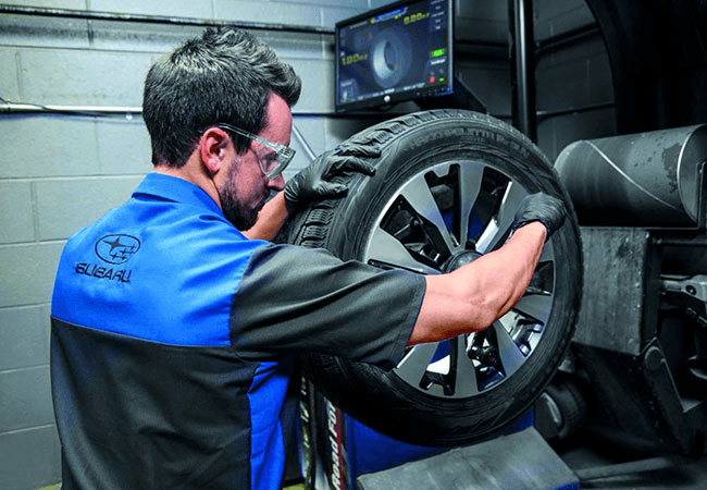 A service technician, wearing a blue and black Subaru uniform and safety glasses, works on a tire and wheel assembly using a tire balancing machine. A monitor in the background displays balancing data.