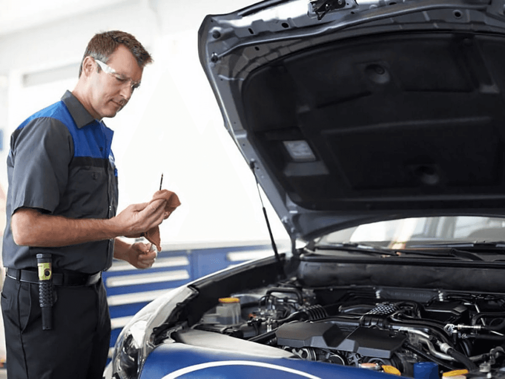 A service technician in a gray and blue uniform and safety glasses is checking the oil dipstick of a vehicle with its hood open in a service bay.