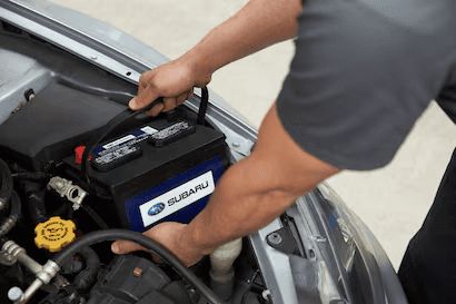 A close-up view of a service technician's hands installing a Subaru-branded car battery in the engine bay of a vehicle.
