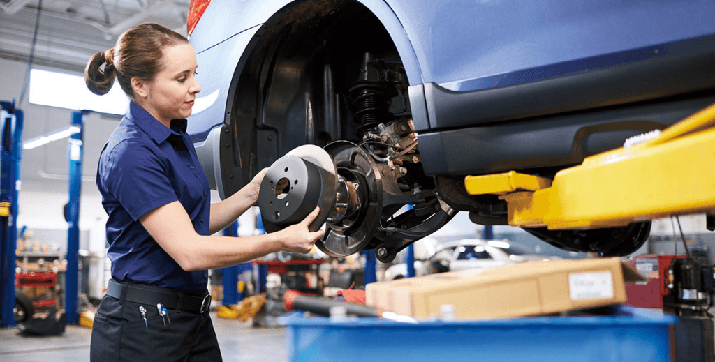 A service technician is working on the brakes of a blue vehicle that is elevated on a lift in an auto repair shop. The technician is holding a brake rotor up to the wheel hub assembly.