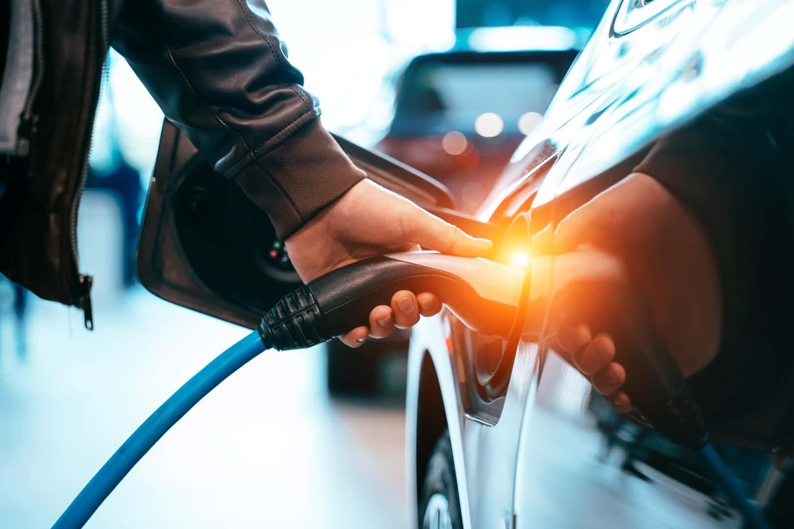 Close-up of a hand plugging an EV charging cable into a vehicle's charge port, with a warm amber glow at the connection point.