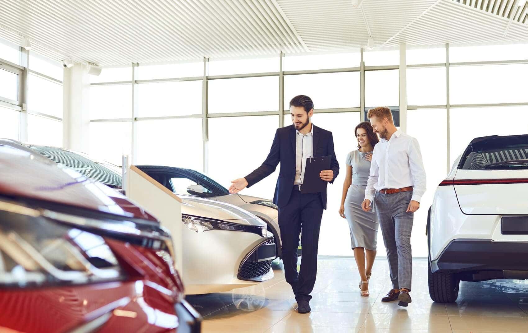 A suited sales consultant walking a smiling couple through a bright, modern dealership showroom, gesturing toward a sedan on the floor