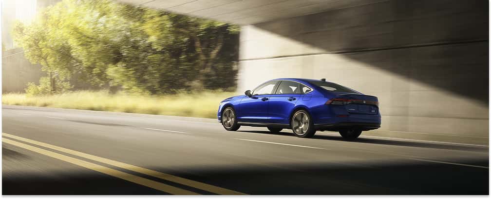 Blue 2026 Honda Accord sedan driving under a concrete overpass, viewed from the rear three-quarter angle.